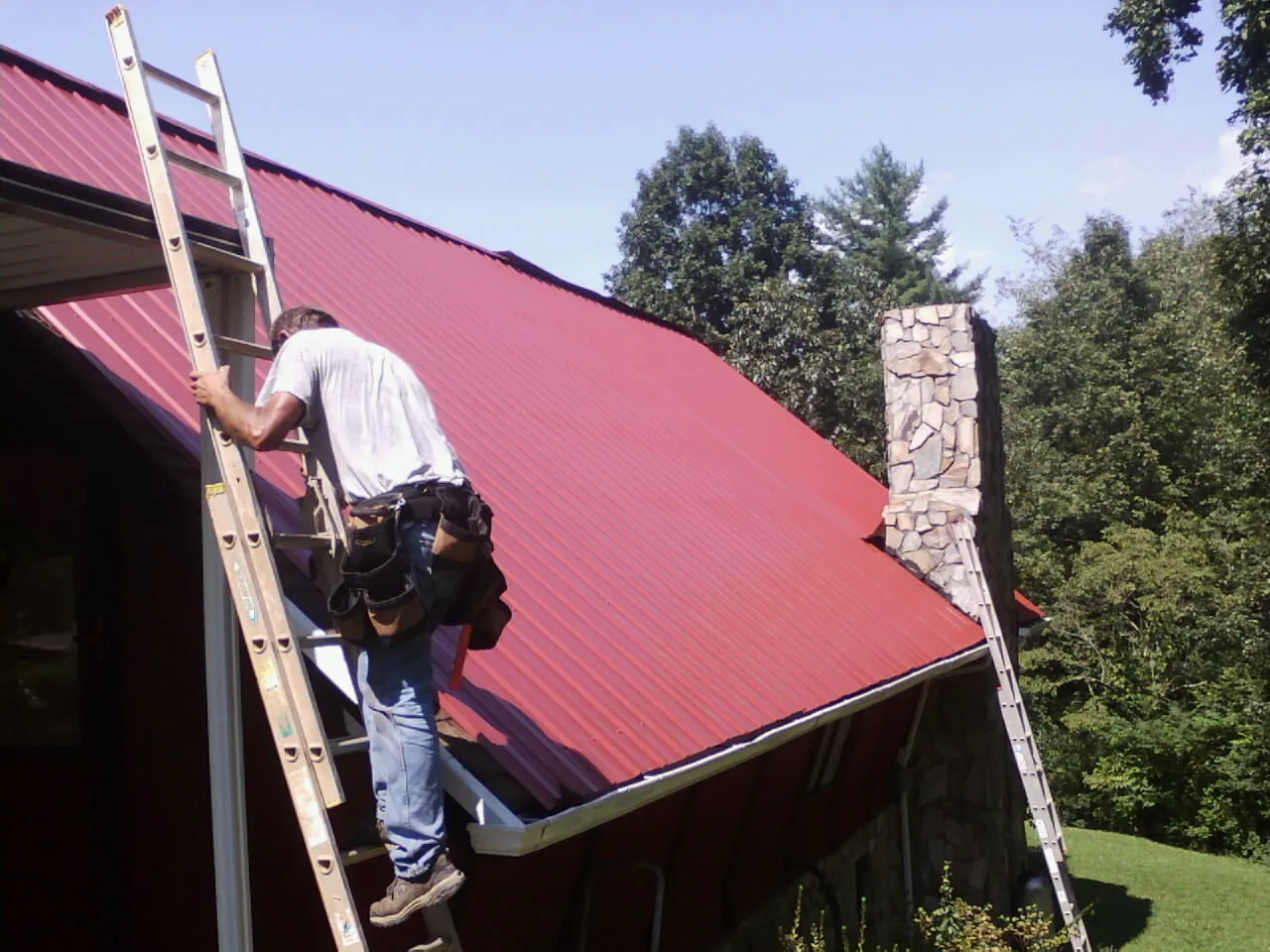 Roofer on a ladder beside a red metal roof and stone chimney — Soffit & Fascia Repair by Roofing Brothers in Cades